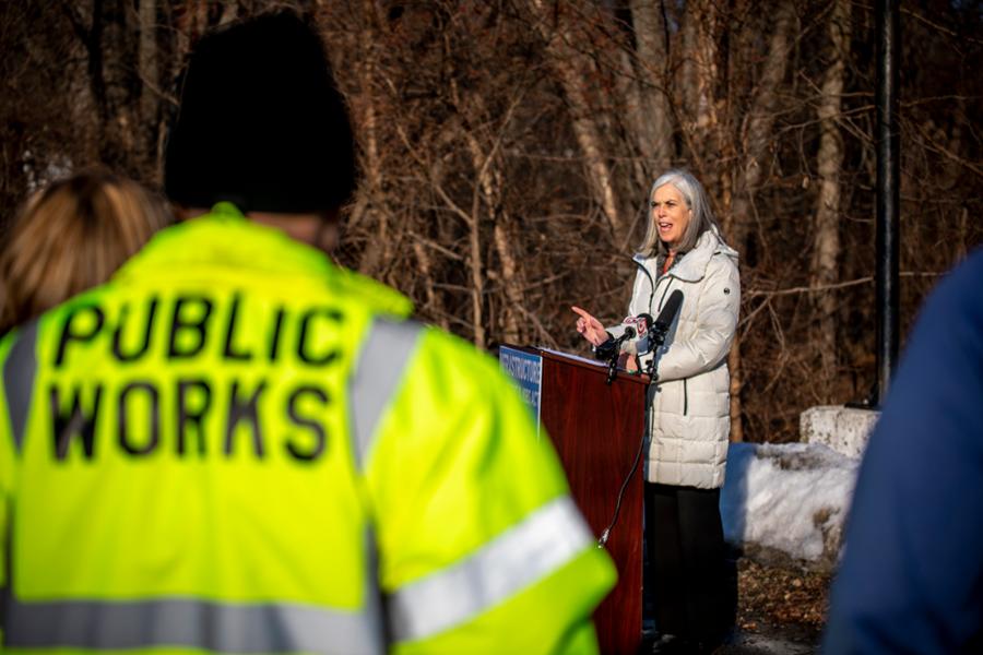 Rep. Clark speaks at a podium in front of trees. In the foreground is a man wearing a reflective coat that says Public Works.