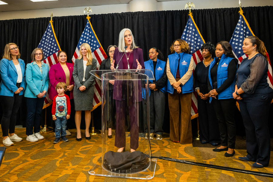 Congresswoman Clark speaks at a podium while surrounded by other women members of Congress. Rep. Clark is wearing is a purple outfit and the podium is translucent.
