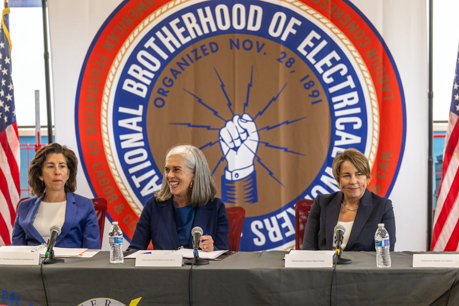 Rep. Clark is sitting at a conference table, flanked by Govenor Maura Healy and Secretary of Commerce Gina Raimonodo. They are in front of a large painting of the local IBEW logo.