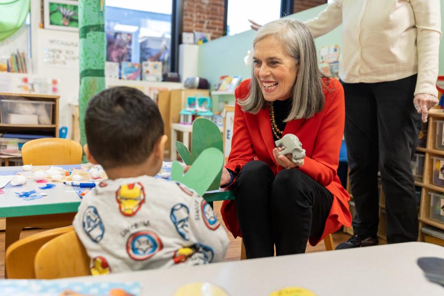 Rep. Clark sits at a kids table with a little boy and holds his toys while laughing in Waltham.